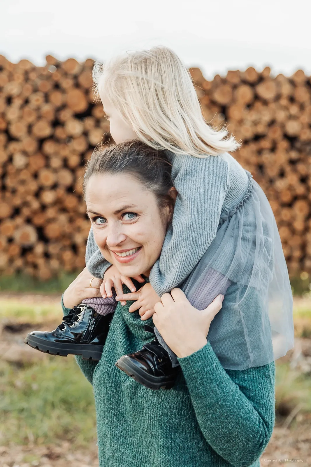 Mama trägt Kind auf dem Rücken vor einem Holzstapel